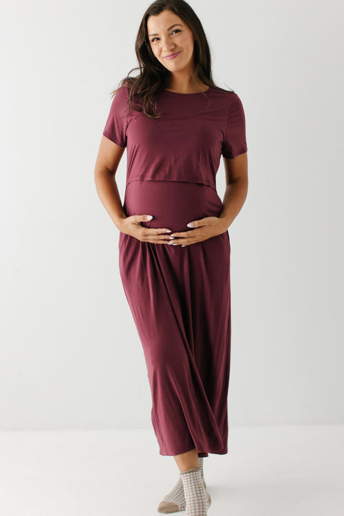 A pregnant woman wearing a short-sleeve, crew-neck maternity dress in a deep burgundy color, standing against a plain white studio background.